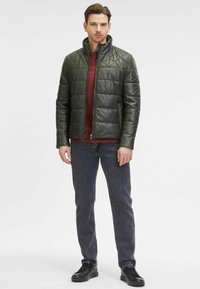 Giacca in pelle trapuntata verde oliva con collo alto, cerniera frontale e design imbottito, indossata sopra una maglietta bordeaux e jeans grigi.