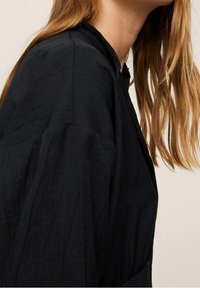 Top in tessuto nero testurizzato con scollatura alta e silhouette rilassata. Le maniche lunghe e i dettagli di cucitura sottili arricchiscono il design.