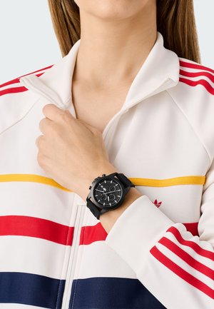 Montre chronographe noire avec un cadran circulaire et trois sous-dials, portée au poignet. Accompagnée d'une veste zippée avec des rayures rouges, jaunes et bleues.