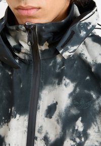 Kurtka o abstrakcyjnym wzorze w czerni i szarości, z wysokim kołnierzem, zamkiem błyskawicznym z przodu oraz wyszywanym logo na lewej piersi.