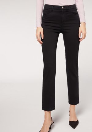 Femme portant un jean noir ajusté et des escarpins slingback noirs à bout pointu, associés à un haut à manches longues rose clair.