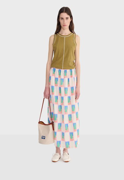 Žena v olivovom topánke bez rukávov, farebne blokovaná dlhá sukňa, biele tenisky, drží béžovú kabelku v tvare vedra s hnedými remienkami.
