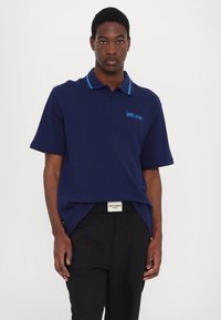 Temno modra polo majica s kontrastnim svetlo modrim ovratnikom, kratkimi rokavi in vezenim logotipom "Just Cavalli" na levem prsnem delu.