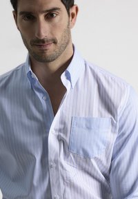 Camisa a rayas en azul claro y blanco, hecha de algodón, presenta un cuello abotonado, un bolsillo frontal y una textura suave.