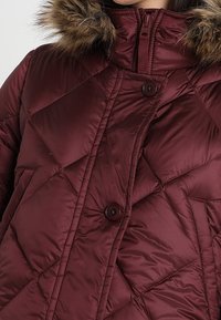 Bordeaux quilted pufferjakke med en fleeceforet hætte, der har en høj krave og to knapper til lukning, som fremhæver en glat tekstur.