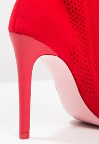 Gros plan sur une chaussure à talons hauts rouges avec un dessus en tissu texturé et un talon aiguille lisse sur un fond blanc.