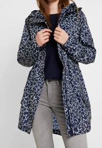 Navy blue rain jacket with white star pattern, adjustable hood, elastic waist, and front zip closure. Lightweight material, mid-length design.