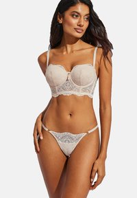 Soutien-gorge en dentelle beige avec armatures et garnitures décoratives ; string assorti avec fines bretelles et détails floraux. Texture douce et design féminin.