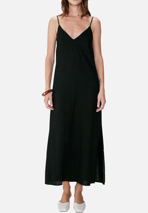 Kvinde iført en lang sort ærmeløs kjole med tynde stropper, hvide spidse sko og et brunt armbånd på håndleddet.