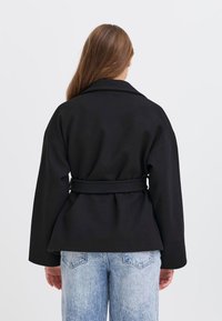 Donna con lunghi capelli castani che indossa un giubbotto nero con cintura e jeans chiari strappati, di spalle contro uno sfondo neutro.