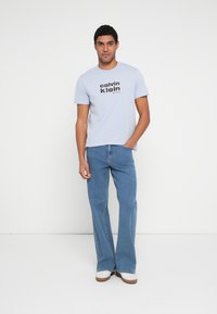 Hele sinine puuvillane t-särk musta "calvin klein" logoga, kombineeritud lehvikujalga siniste teksapükstega. Modell seisab ühevärvilise valge tausta ees.