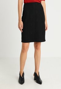 Jupe crayon noire en tricot avec un motif texturé, de longueur genou et une forme subtile. Portée avec un haut rouge et des chaussures à talons noires.