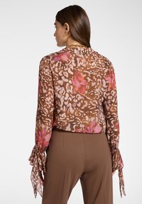 Blusa marrone a motivo con dettagli floreali rosa, maniche lunghe con polsini arricciati, indossata con pantaloni marroni. Tessuto leggero e trasparente.
