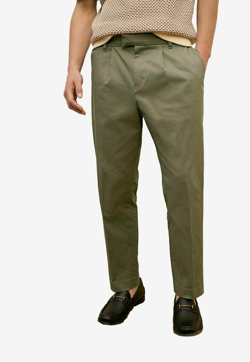 Olivengrønne skræddersyede bukser med en glat stofoverflade, med frontfolder og sidelommer, parret med sorte slip-on sko.