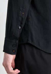 Svart långärmad skjorta med knappmanschetter, orange accenttråd och en något rundad nederkant. Tyget verkar mjukt och strukturerat.