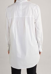 Blusa bianca a maniche lunghe con scollo arruffato, chiusura con bottoni sul retro e dettagli plissettati, che si estende sotto i fianchi per un fit rilassato.