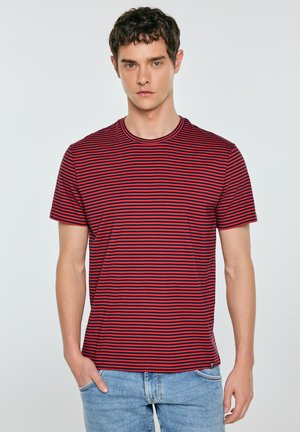 Młody mężczyzna z krótkimi kręconymi włosami, ubrany w czerwono-czarną koszulkę w paski z krótkim rękawem i jasnoniebieskie dżinsy, stoi z jedną ręką w kieszeni.