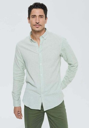 Chemise à carreaux verts boutonnée en tissu léger, avec un col classique et des manches longues. Portée avec un pantalon vert foncé.