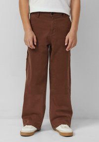 Brązowe szerokie spodnie cargo wykonane z trwałej tkaniny, wyposażone w boczne kieszenie, zapięcie na guziki z przodu i luźny krój. Noszone z białymi tenisówkami.