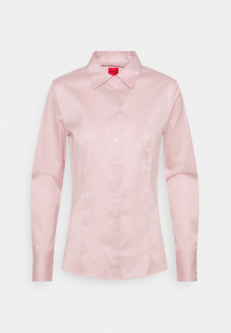 Chemise rose pâle à boutons, fabriquée en tissu lisse et brillant. Présente un col classique et des manches longues avec une silhouette ajustée. Étiquette logo à l'intérieur du col.