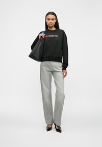 Černá mikina s růžovým textem, šedé rovné džíny a černé lodičky s špičatou špičkou. Nese černou kabelku přes rameno. Jednoduché pozadí.