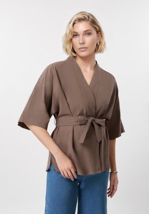 Kobieta z krótkimi blond włosami, ubrana w brązową bluzkę o szerokich rękawach z wiązanym paskiem, w połączeniu z niebieskimi dżinsami, stojąca na białym tle.