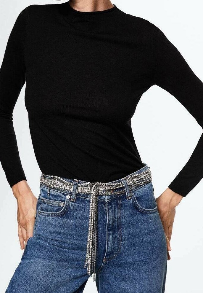 Haut noir à manches longues associé à un jean en denim bleu. La ceinture présente un motif de strass argenté avec un détail de chaîne pendante.