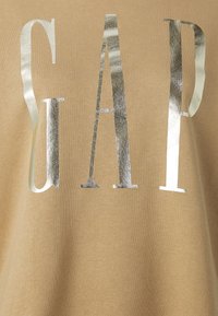 Camiseta de algodón color caqui con grandes letras metálicas plateadas "GAP". Textura suave con un corte relajado y un diseño minimalista.