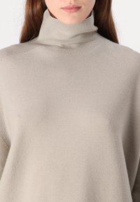 Beige neulepusero, joka on valmistettu pehmeästä neulosmateriaalista. Ominaisuuksina rento istuvuus, ribattua tekstuuria, eikä näkyviä kuvioita tai varusteita.