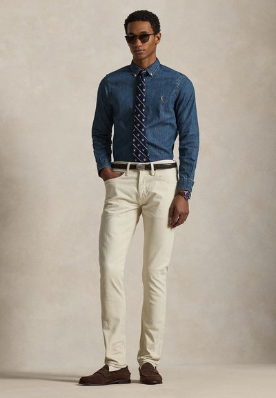 Uomo con occhiali da sole scuri, camicia di jeans con bottoni, cravatta blu navy, pantaloni color avorio, cintura e scarpe marroni, in piedi su uno sfondo beige uniforme.
