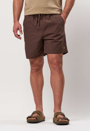 Mies, jolla on päällään ruskeat narusäätöiset shortsit ja ruskeat sandaalit, seisoo käsi taskussa yksiväristä taustaa vasten.