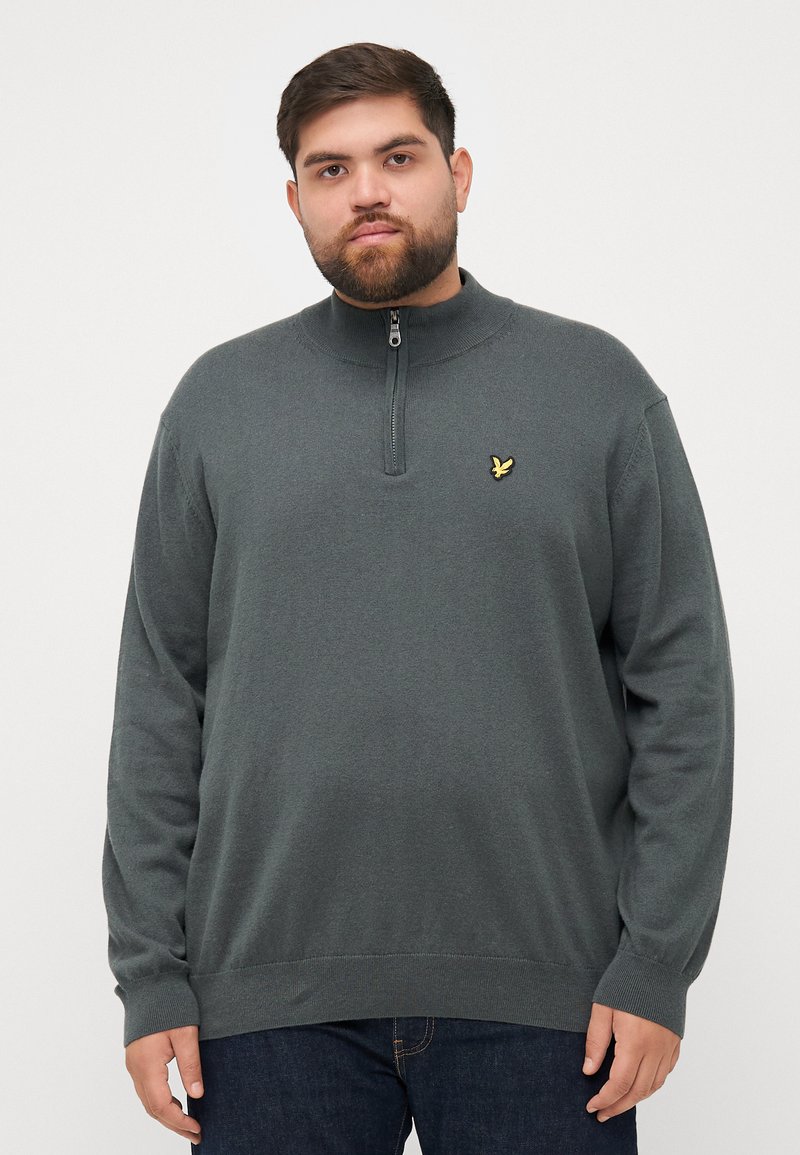 Pullover gris foncé avec un col zippé mi-hauteur, des poignets et un ourlet côtelés, avec un petit logo jaune sur la poitrine gauche. Texture de tissu douce.