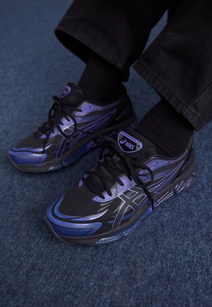 Des chaussures de sport noires et violettes présentent un dessus en mesh, une semelle rembourrée et un logo bien en évidence sur la languette, avec des lacets et des accents noirs.