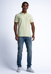 Ljusgul polo med krage och logga på vänster bröst; kombinerad med blå jeans och vita sneakers. Enkelt mönster, avslappnad passform.