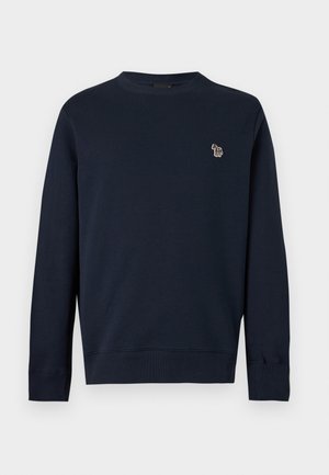 Junger Mann steht mit den Händen in den Taschen, trägt ein marineblaues Sweatshirt, graue Jogginghosen und weiße Sneakers vor einem einfarbigen weißen Hintergrund.