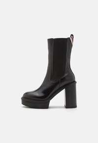 Zwarte enkelboot met een glanzend leren bovenkant, elastische zijpanelen en een chunky, gestructureerde plateau hak. Heeft een lus aan de achterkant.