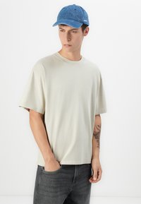 Lys beige kortærmet T-shirt med en afslappet pasform, parret med en blå denimkasket med et frontlogo og grå jeans.