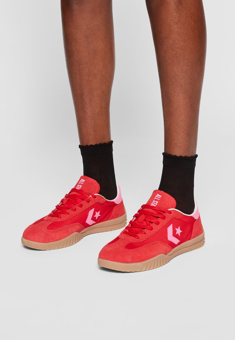 Czerwone zamszowe trampki z różowym akcentem na pięcie, z czerwonymi sznurowadłami, gumową podeszwą i logotypem gwiazdy z boku. Czarne skarpetki za kostkę.