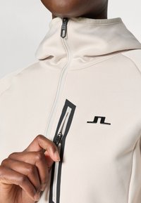 Lichtbeige zip-up jas gemaakt van een gladde stof. Beschikt over een hoge kraag, zwarte rits en een zwart logo op de voorkant.