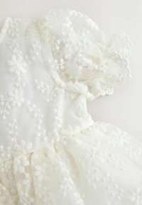 Fehér csipkeruha virágos hímzéssel, áttetsző anyaggal, pufós ujjal és fodros szélű, ráncolt szoknyával.
