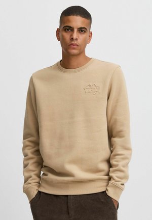 Beige sweatshirt med rund hals og lange ærmer. Har et præget "ENJOY" logo og et tekstureret stof. Paires med mørkebrune bukser.