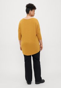 Mustard yellow oversized top with a rounded hem, three-quarter sleeves, and a relaxed fit, paired with black wide-leg trousers.