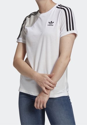 Osoba ubrana w białą koszulkę Adidas z czarnymi paskami na rękawach i logo na piersi, w połączeniu z niebieskimi dżinsami, ręce skrzyżowane na talii.