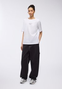 T-shirt bianca a maniche corte con una stampa sottile, abbinata a pantaloni neri larghi con cuciture a contrasto e tasche. Sneakers argentate.