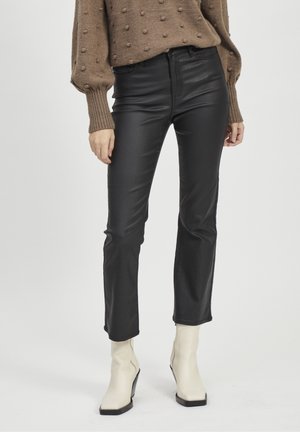 Femme portant un pantalon en cuir noir, un pull en tricot beige avec des points texturés et des bottines blanches, debout sur un fond blanc.