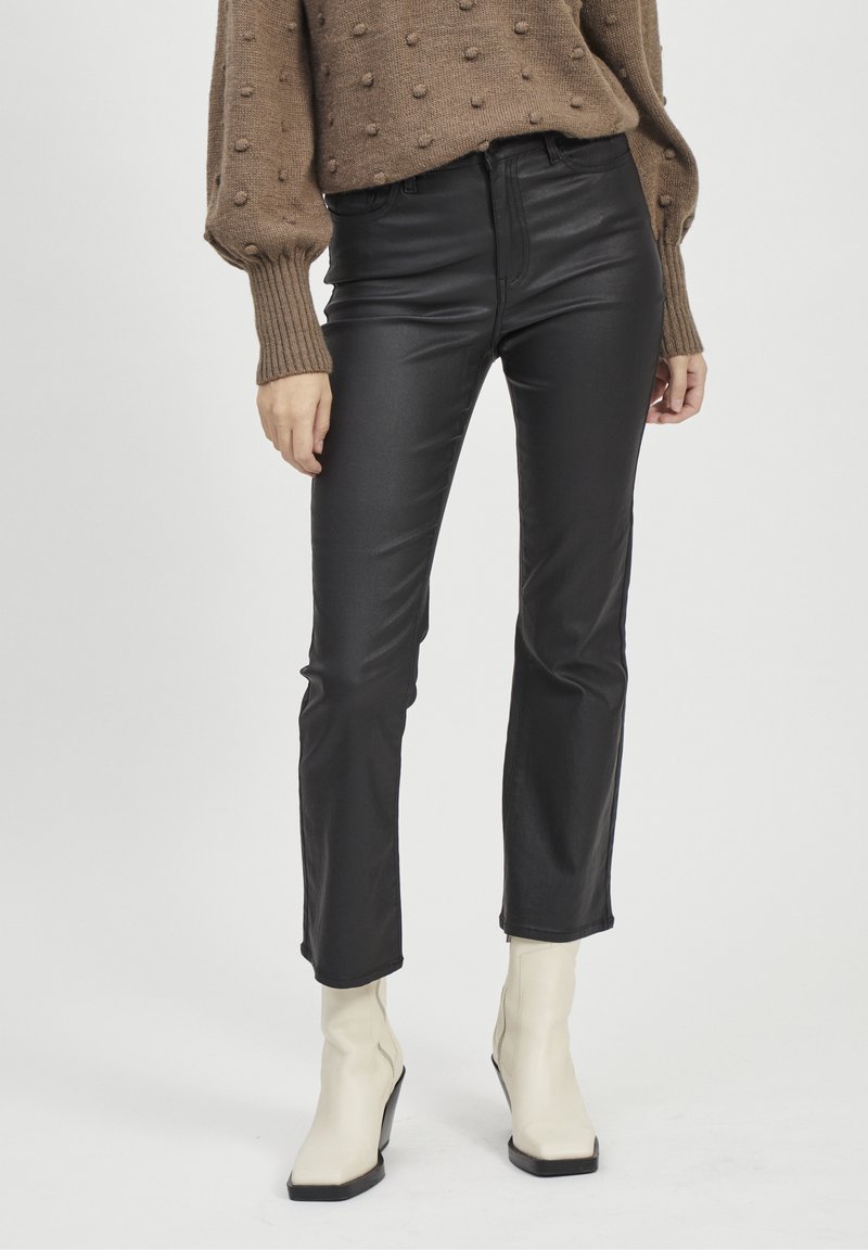 Femme portant un pantalon en cuir noir, un pull en tricot beige avec des points texturés et des bottines blanches, debout sur un fond blanc.