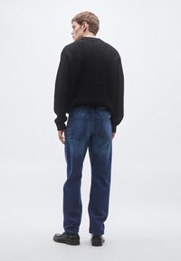 Mustast paksudest kootud kampsun ümarate kaelustega, kombineeritud tumedatest sinistest sirge lõikega teksapükstega ja mustade pidulike jalanõudega. Tagumine vaade.