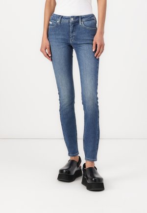 Jeans ajustados de denim, lavado azul medio, con cinco bolsillos, presillas para cinturón y un leve desgaste. Llevados con zapatos de plataforma negros.