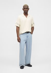 Homme aux cheveux blonds courts portant une chemise crème à manches courtes boutonnée avec des motifs, un jean large bleu clair, une ceinture noire et des chaussures noires.