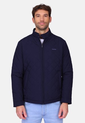 Homme portant une veste matelassée bleu marine zippée et un jean bleu clair, debout devant un fond clair uni.